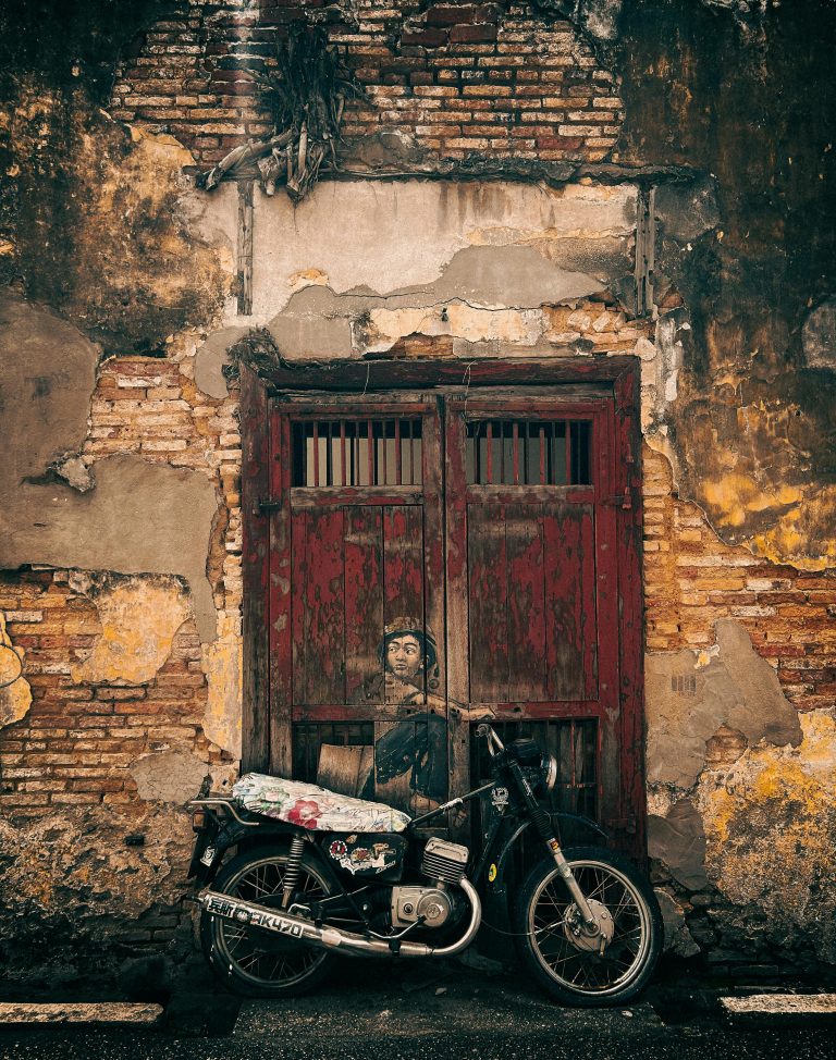 A vintage motorcycle parked against a rustic, weathered wall in George Town, Penang, Malaysia. Urban art scene.